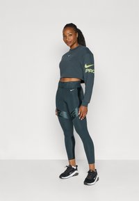 Tamno siva skraćena dukserica s zelenim Nike logotipom, uparena s tamno zelenim tajicama s Nike trakama i crnim tenisicama.