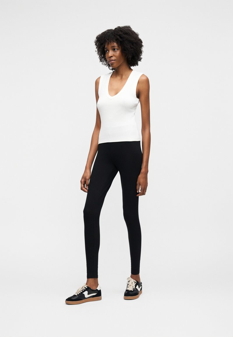 Donna con capelli ricci che indossa una canotta bianca, leggings neri e scarpe da ginnastica bianche e nere, in piedi davanti a uno sfondo semplice.