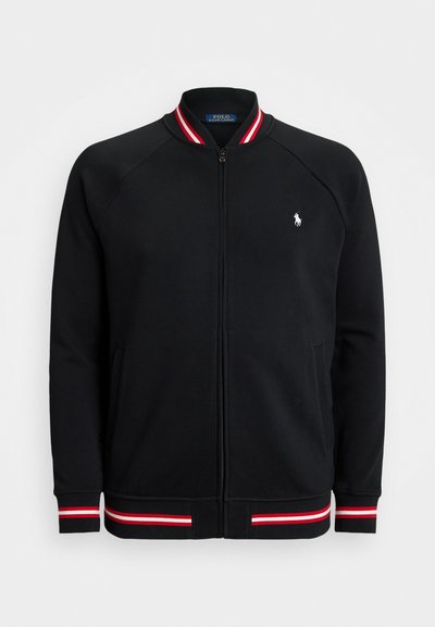 Musta vetoketjullinen takki, joka on valmistettu pehmeästä kankaasta. Takissa on ribbikaarroke, jossa on punaiset ja valkoiset raidat, sekä pieni valkoinen logo rinnassa.