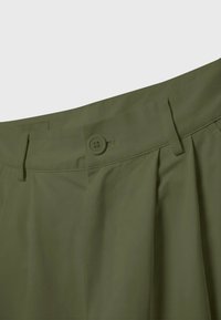 Gros plan sur la ceinture d’un pantalon vert olive avec fermeture par bouton et passants de ceinture sur un fond uni.