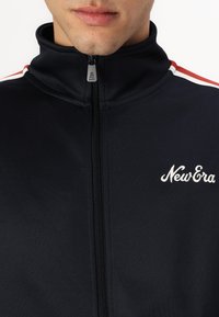 Ναυτικό μπουφάν με φερμουάρ που έχει ψηλό γιακά, λευκές και κόκκινες ρίγες στους ώμους και κεντημένο λογότυπο "New Era" στο στήθος.