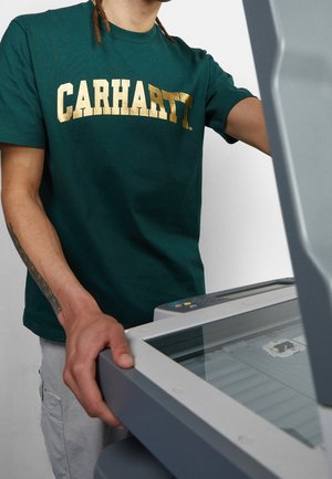 Persoon in een groen Carhartt-T-shirt bedient een grijze vlakbedscanner of kopieerapparaat met deksel open tegen een effen achtergrond.