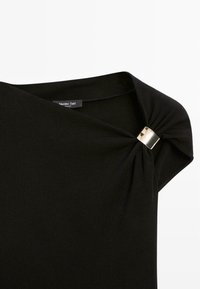 Svart topp tillverkad av slät tyg, med en draperad design som har en vriden knut och en metallisk detalj vid axeln.