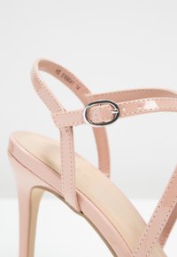 Sandale à talon haut en cuir verni rose avec des fines brides et une petite boucle argentée sur fond blanc.