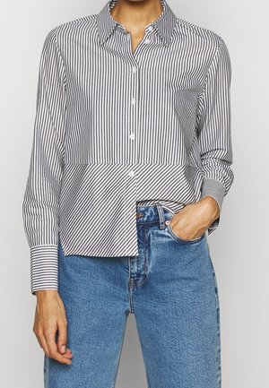 Frau trägt ein schwarz-weiß gestreiftes Hemd mit vertikalen Streifen und diagonalen Streifen im unteren Bereich, kombiniert mit blauen Jeans.