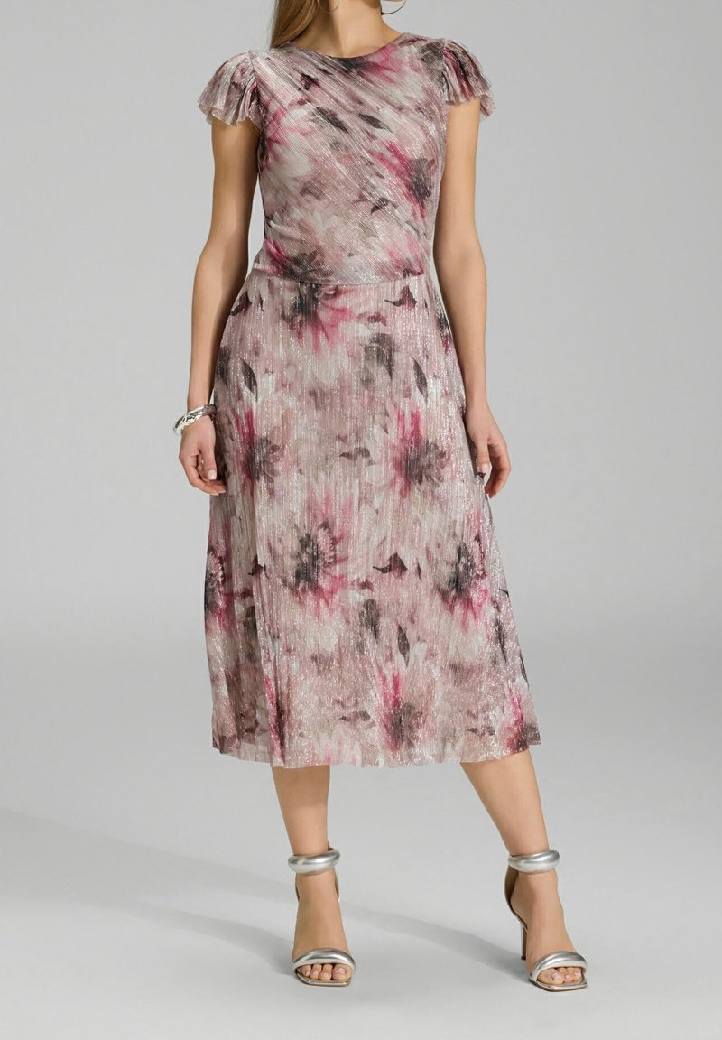 Femme portant une robe midi fleurie rose et grise avec des manches bouffantes courtes et des sandales à talons hauts argentées, debout devant un fond uni.