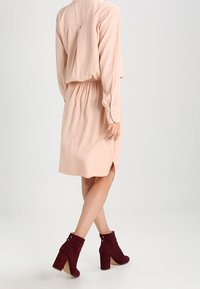 Šaty v broskvové barvě s dlouhými rukávy a elastickým pasem, s skladem na zadní straně. Nosené s burgundskými semišovými kotníkovými botami na širokém podpatku.