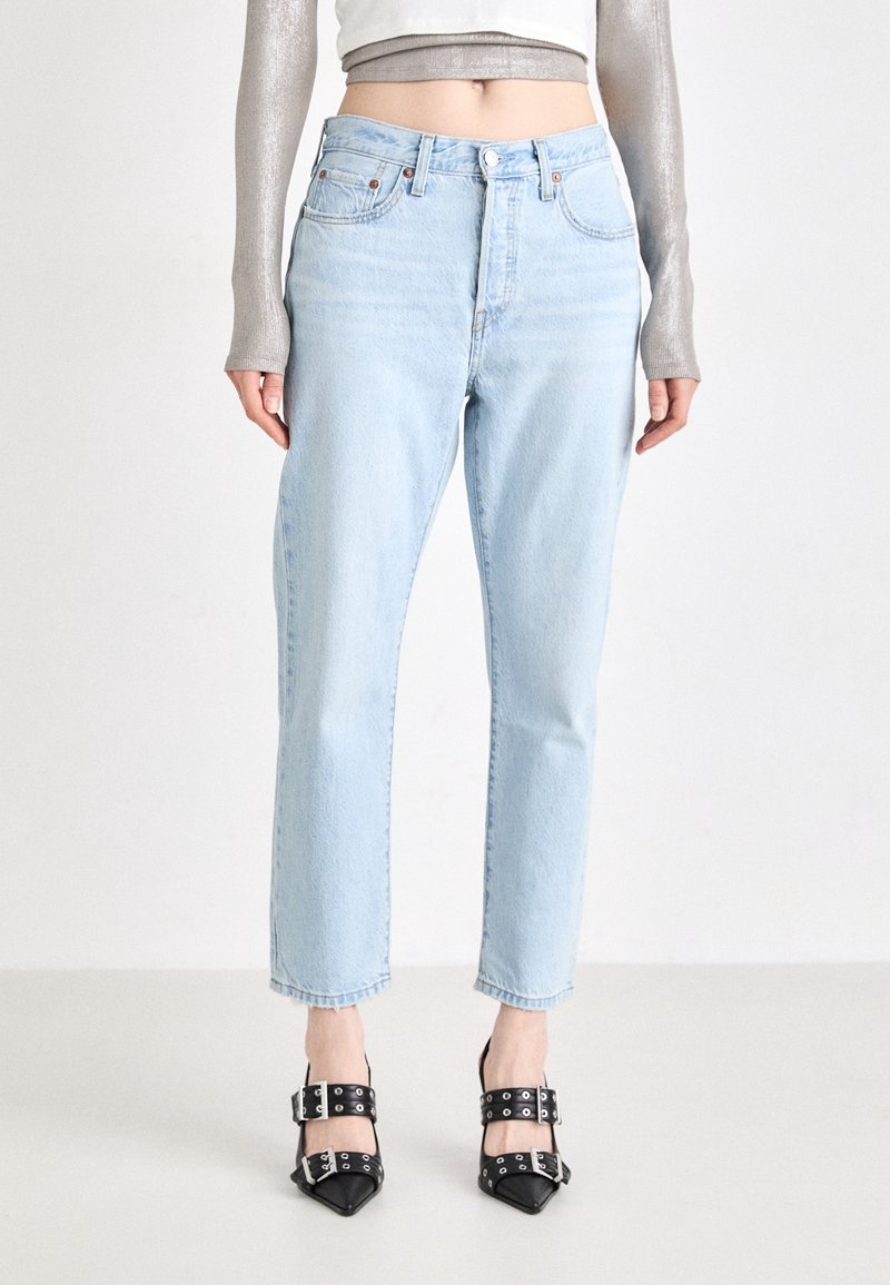Persoon in lichtblauwe high-waist jeans, een cropped langmouw metallic zilveren top en zwarte puntige hakken met dubbele gespen.