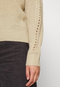 Beige stickad tröja med en texturerad mönster, med hålstickade detaljer längs ärmen. Ribbad mudd och en figursydd silhuett.
