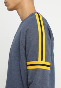 Felpa blu con maniche lunghe, con dettagli a strisce gialle e nere sulle spalle. Tessuto morbido e vestibilità comoda.