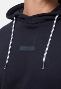 Navy blauwe hoodie met een gestructureerde stof, witte koorden met zwarte accenten en een rechthoekig logo op de borst.