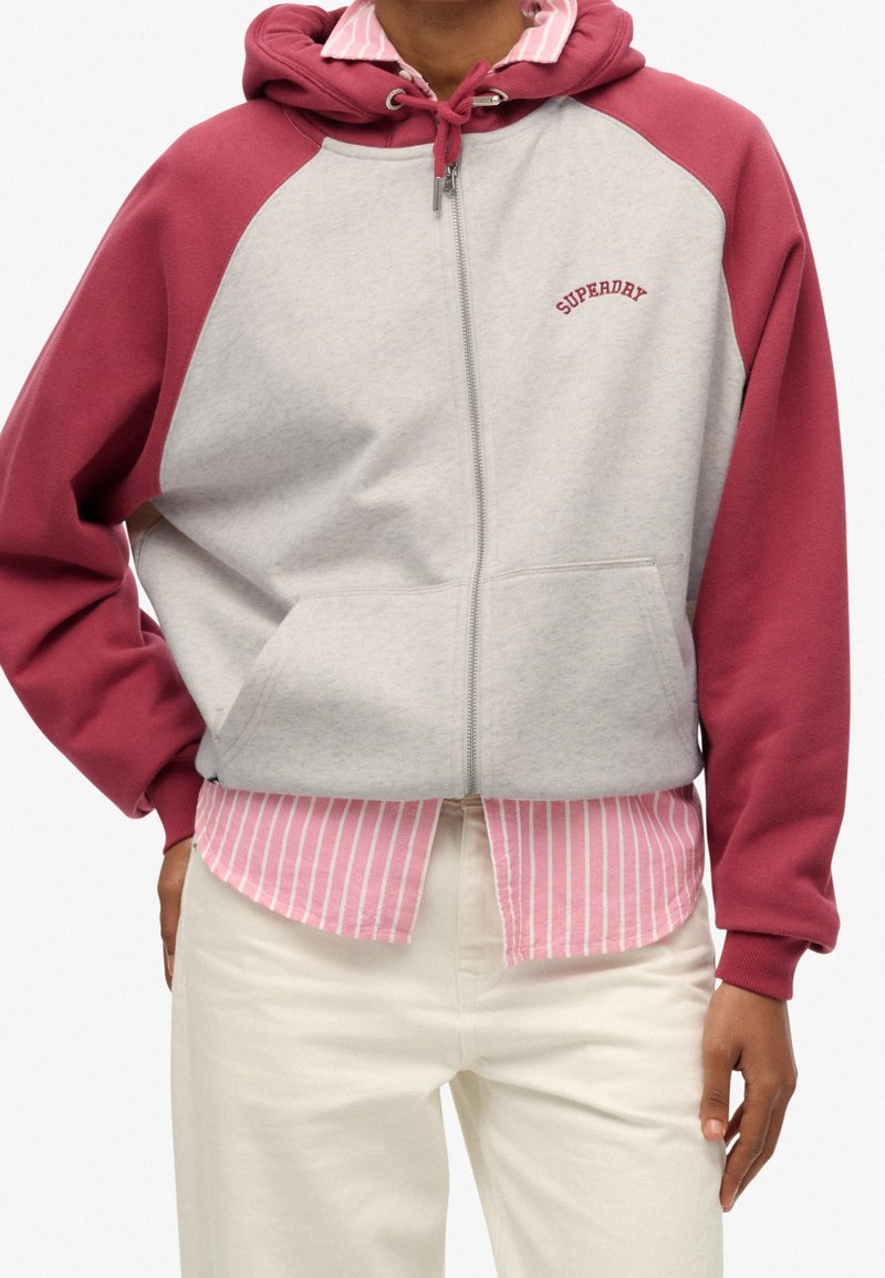 Personne portant un sweat à capuche zippé avec corps gris, manches roses foncées, texte « SUPERDRY » sur la poitrine, par-dessus une chemise à rayures roses et blanches et un pantalon blanc.