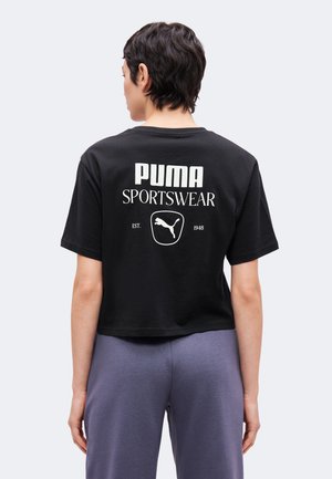 Osoba s krátkymi tmavými vlasmi, ktorá má na sebe čierne krátke športové tričko Puma a sivé nohavice, viditeľná zozadu.