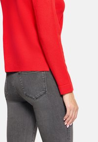 Czerwony sweter z dzianiny o zaokrąglonym dole i długich rękawach, w połączeniu z ciemnoszarymi dżinsami z tylnymi kieszeniami i dopasowanym krojem.
