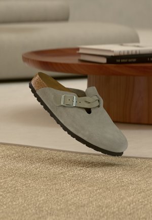 Világosszürke velúr klumpa csatos pánttal, amely egy modern nappali környezetben lebeg egy bézs szövött szőnyeg fölött.