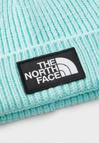 Berretto lavorato a maglia di colore azzurro chiaro con texture a coste e un'etichetta rettangolare nera con testo e logo bianchi.