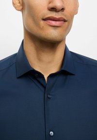 Navyblaues Hemd mit einem dezenten Diamantenmuster, geknöpftem Kragen und zwei sichtbaren Knöpfen auf der Knopfleiste, das einen strukturierten Stoff zeigt.