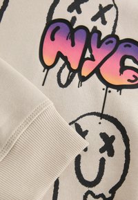 Beżowy sweatshirt z prążkowanym mankietem, z czarną grafiką uśmiechu i kolorową metką "NYC" w gradientowych odcieniach różu i pomarańczy.