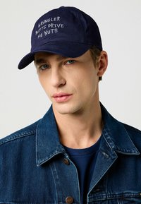 De marinepet heeft witte geborduurde tekst "WRANGLER BUTTS DRIVE ME NUTS." Het model draagt een spijkerjack met daaronder een blauwe shirt.