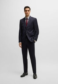 Un costume bleu marine en tissu lisse, à simple boutonnage, avec une chemise blanche à motifs et une cravate bordeaux. Des chaussures noires complètent le look.