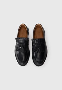 Mocassins en cuir noir avec des franges et des détails à pampilles, doublure intérieure marron, présentés côte à côte sur un fond blanc.