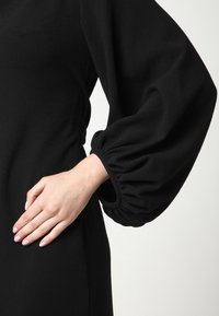 Robe noire en tissu texturé, avec manches bouffantes froncées et silhouette ajustée. Design simple et élégant avec un minimum de détails.