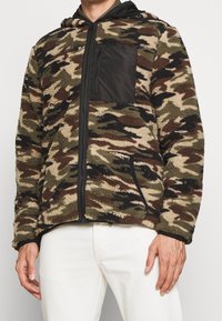 Maskáčová fleecová mikina s kapucí, na hrudi kapsa na zip, černé detaily a přední kapsy. Vyrobena z texturovaného, teplého materiálu v zemitých tónech.