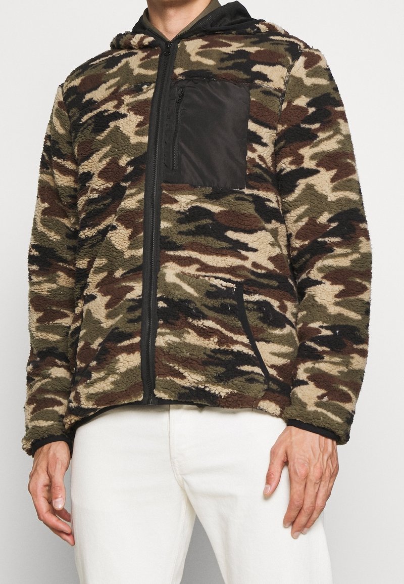 Maskáčová fleecová mikina s kapucí, na hrudi kapsa na zip, černé detaily a přední kapsy. Vyrobena z texturovaného, teplého materiálu v zemitých tónech.