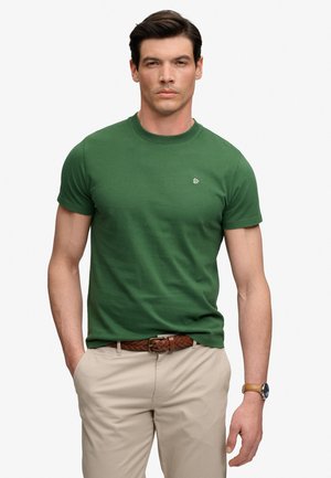 Mann trägt ein grünes kurzärmeliges T-Shirt mit Rundhalsausschnitt, beige Hose mit gewebtem braunem Gürtel und Armbanduhr, steht vor weißem Hintergrund.
