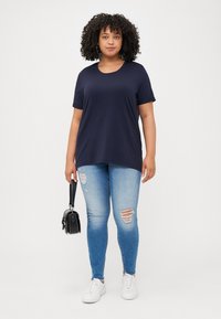 Curly-haired person wearing a navy t-shirt and ripped light blue jeans, holding a small black handbag, stands against a neutral background.