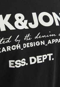 T-shirt in cotone nero con testo bianco bold. Il testo include il nome del marchio e frasi come "ricerca", "design" e "ess. dept." in diversi caratteri.