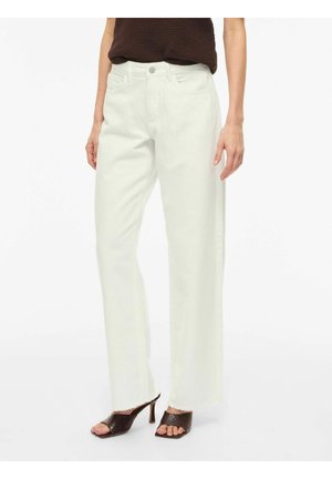 Kobieta ubrana w białe, szerokie jeansy z wysokim stanem i postrzępionymi brzegami oraz brązowe sandały na obcasie, połączone z ciemnym, bezrękawnym topem.