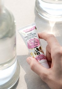 Korres Damask Rose-mask i en vit tub. Har rosa blommönster och guldfärgade detaljer. Hand som håller tuben nära ett glas vatten på en ljus yta.