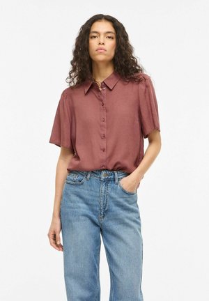 Sieviete ar cirtainiem matiem, valkājot īsroku krekla brūngani rudu blūzi, kas iestrādāta augsti jostas gaismās zilās džinsa biksēs, viena roka kabatā.