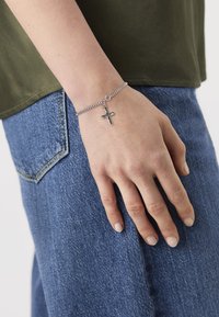 Sølv kjedearmbånd med en liten korsettsjarm, båret på en hånd. Armbåndet har en polert overflate; hånden hviler på denimstoff.
