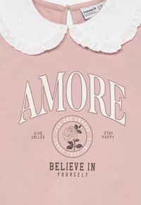 Rosa bomullstop med en hvit volangkrage, fet tekst "AMORE" og blomster grafikk. Inkluderer tekst: "Du er nok, del smilet ditt."