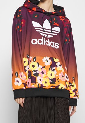 Bluza z kapturem z logo Adidas, tło gradientowe od fioletu do pomarańczowego oraz żółte, różowe i czerwone wzory kwiatowe, noszona przez osobę w czarnej plisowanej spódnicy.