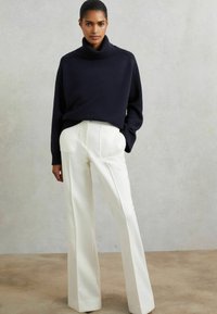 Pull à col roulé bleu marine avec des poignets côtelés associé à un pantalon blanc ajusté à taille haute et jambe large. Textures lisses, design minimaliste.