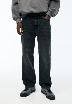 Person trägt ausgewaschene schwarze Jeans mit weitem Bein, grauen Hoodie und grau-schwarze Sneakers, steht mit einer Hand in der Tasche vor einem einfarbigen Hintergrund.