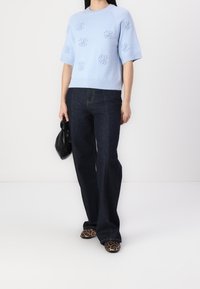 Hellblauer Strickpullover mit bestickten Blütendetails, kombiniert mit dunkelblauen Schlagjeans und Leopardenmuster-Schuhen, hält eine schwarze Tasche.