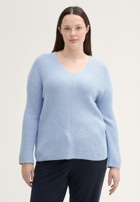 Jasnoniebieski sweter dzianinowy z dekoltem w serek, z teksturowanym wzorem w zygzak. Długie rękawy i luźny krój, w połączeniu z ciemnymi spodniami.