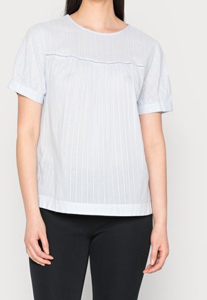 Woman wearing a short-sleeve, white textured blouse with subtle vertical stripes and black pants against a plain background.