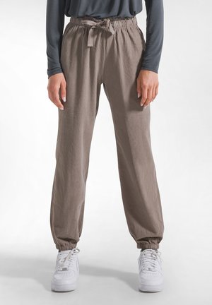 Osoba nosząca taupe joggery z elastycznym pasem, sznurkiem i elastycznymi mankietami, w połączeniu z białymi sneakersami i ciemnoszarą koszulką z długim rękawem.