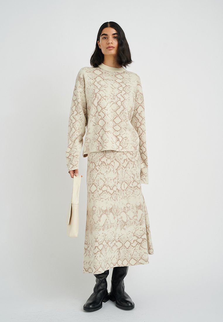 Beiges Sweater- und Rock-Set mit einem Schlangenprint-Muster, langen Ärmeln und einer lockeren Passform. Kombiniert mit schwarzen kniehohen Stiefeln und einer hellen Tasche.