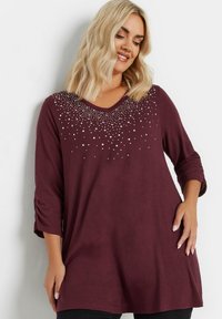 Burgundy pitkähiha tunika V-pääntiellä, jossa on monimutkainen hopean värinen helmikoristelua kauluksessa ja käännetyt hihansuut. Pehmeä, teksturoitu kangas.