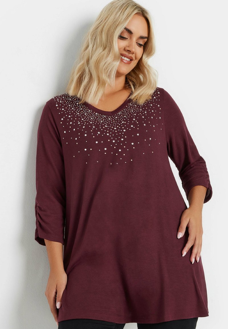 Burgundy pitkähiha tunika V-pääntiellä, jossa on monimutkainen hopean värinen helmikoristelua kauluksessa ja käännetyt hihansuut. Pehmeä, teksturoitu kangas.