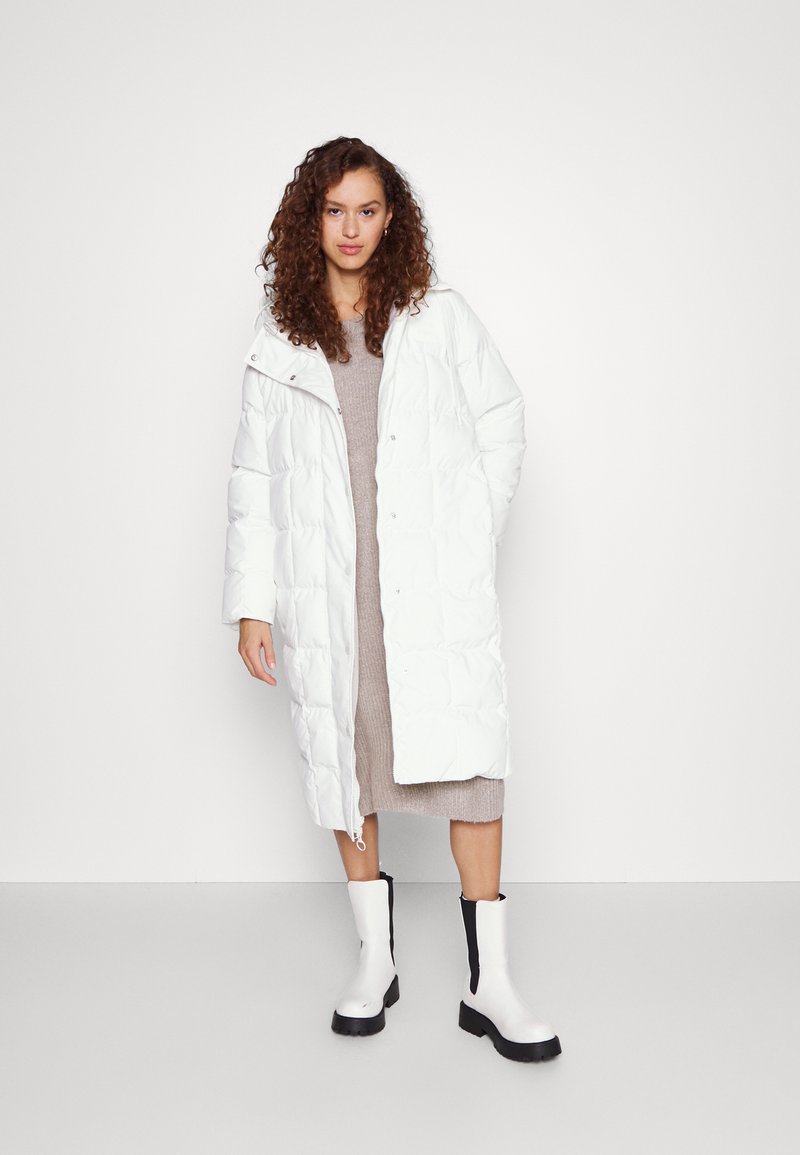 Femme avec des cheveux bouclés portant un long manteau en duvet blanc, une robe en tricot beige et des bottes blanches avec des semelles noires, se tenant devant un fond blanc uni.