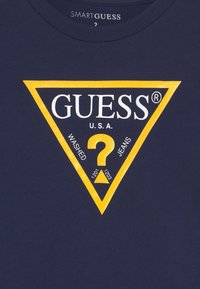 Tejido azul marino con un triángulo amarillo que presenta el texto "GUESS U.S.A.", un signo de interrogación amarillo y las palabras "WASHED" y "JEANS."
