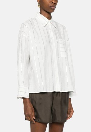 Donna che indossa una camicetta bianca con bottoni, righe verticali di paillettes e taschino sul petto, abbinata a pantaloncini in raso marrone scuro.