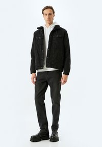 Giubbotto di jeans nero con colletto in shearling, abbinato a una felpa bianca, jeans neri e stivali neri chunky, su uno sfondo bianco.